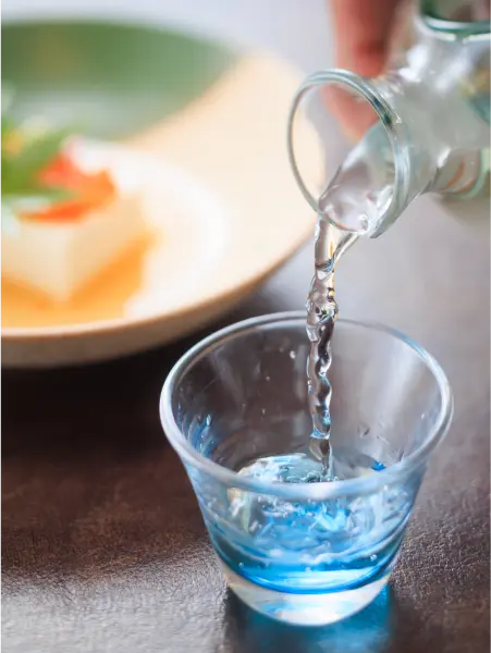 青いガラスの器に日本酒を注ぐ。奥には豆腐料理。