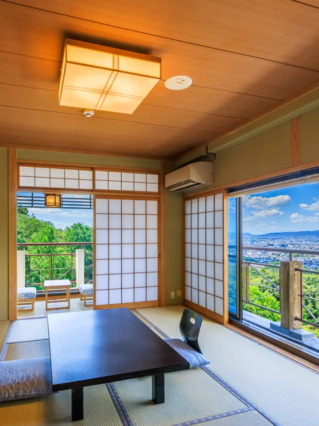 畳の部屋に置かれた黒いテーブルと座椅子。障子と大きな窓からは、青い空と広がる街並みが見えます。