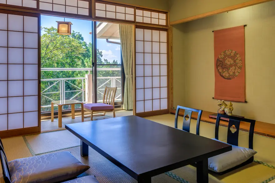 障子で仕切られた和室。低いテーブルと座椅子が置かれ、窓からは青々とした木々が見えます。