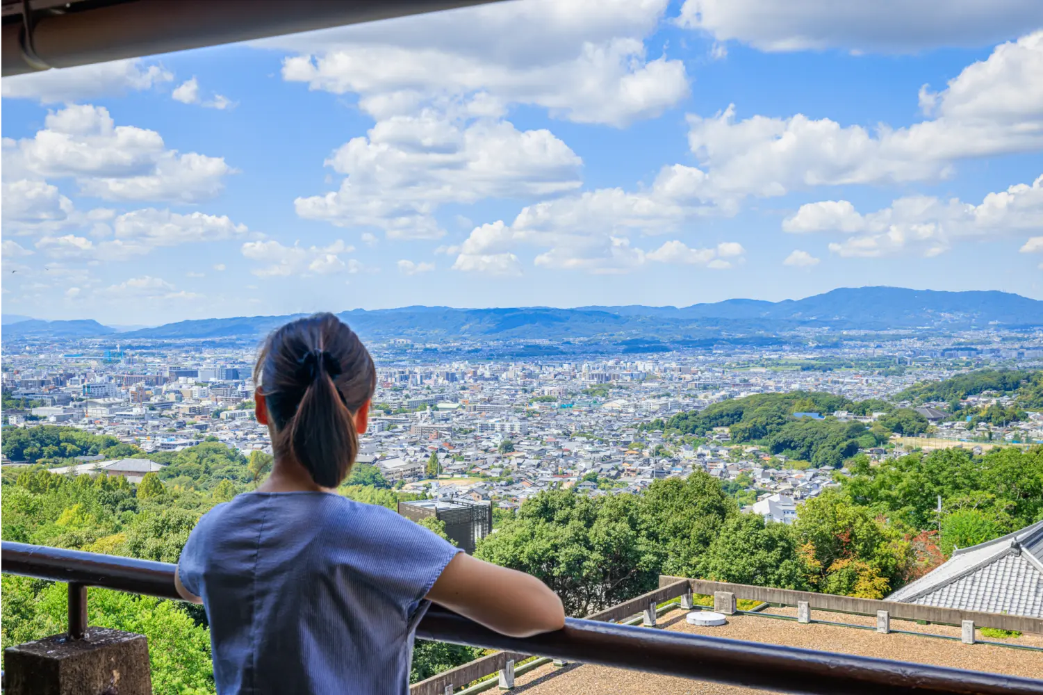 晴れた日にバルコニーから街並みを眺めている女性の後ろ姿。遠くには山々が見えます。