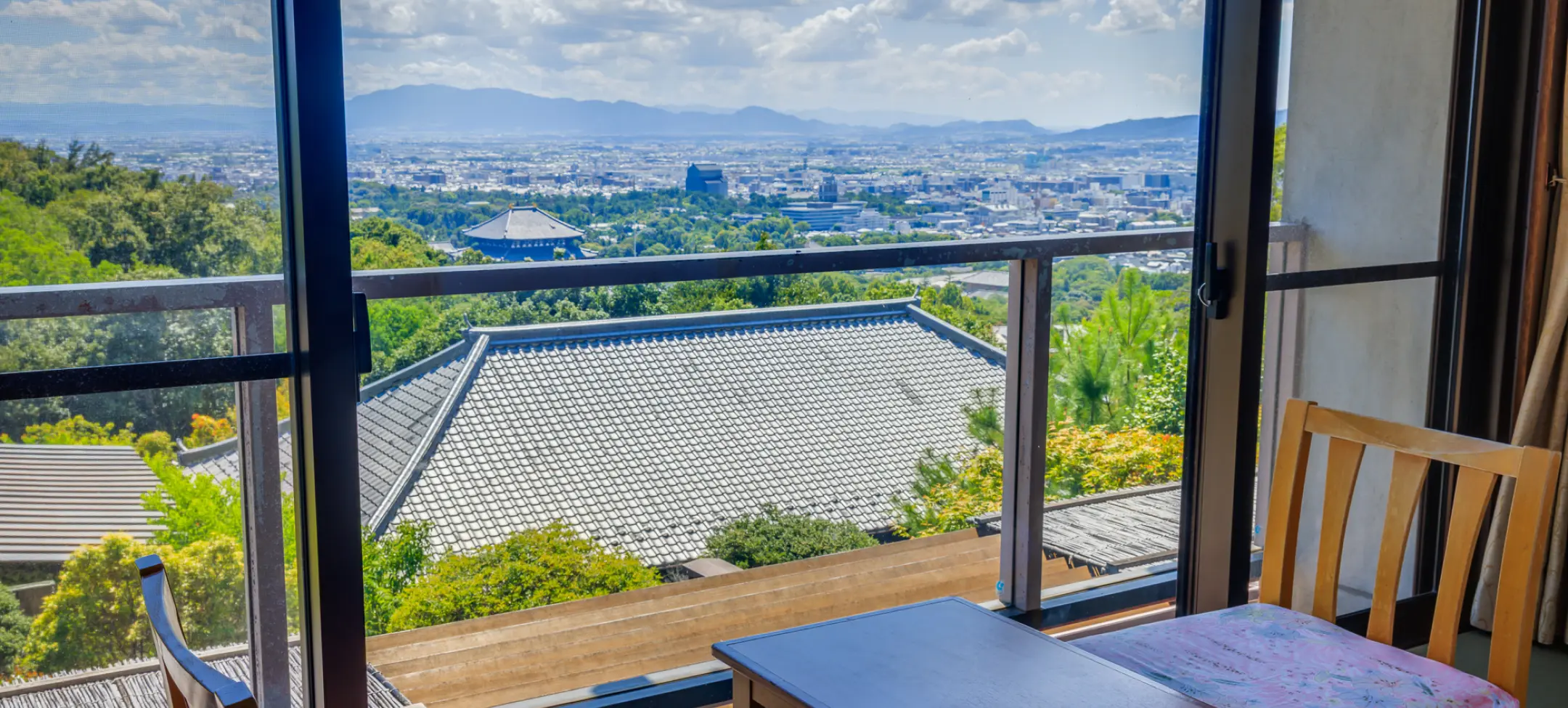 窓際の客室から昼間に見た景色。手前に木製の椅子と小さなテーブルがあり、窓越しには和風の屋根と、その先に広がる山と市街地が見える。