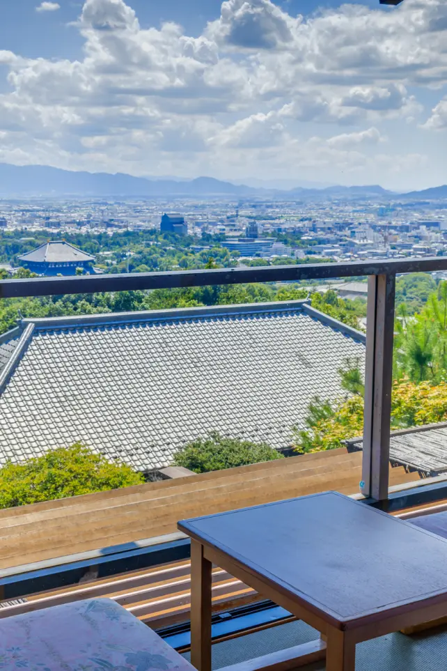 窓際の客室から昼間に見た景色。手前に木製の椅子と小さなテーブルがあり、窓越しには和風の屋根と、その先に広がる山と市街地が見える。
