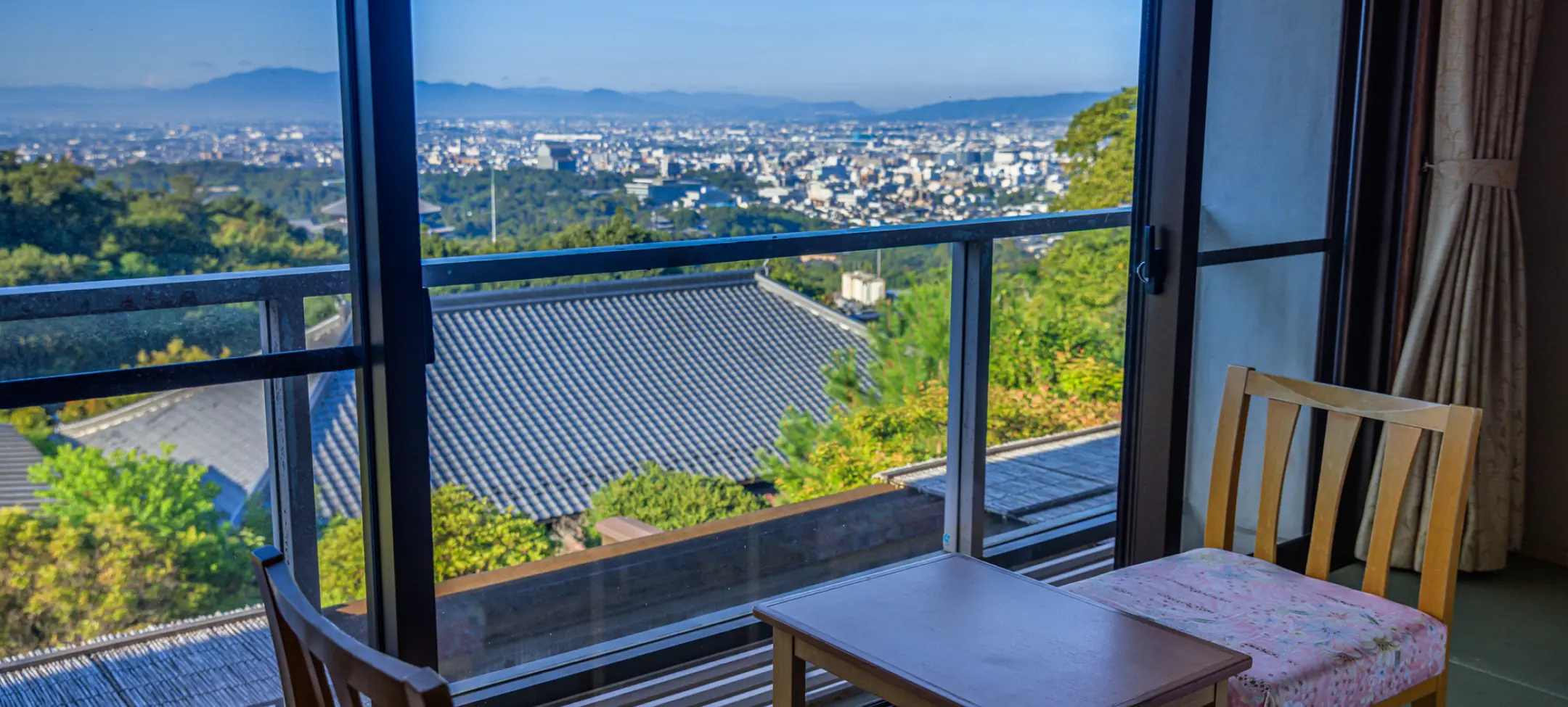 窓際の客室から夕暮れ時に見た景色。手前に木製の椅子と小さなテーブルがあり、窓越しには和風の屋根と、遠方に薄明かりに照らされた市街地が広がる。