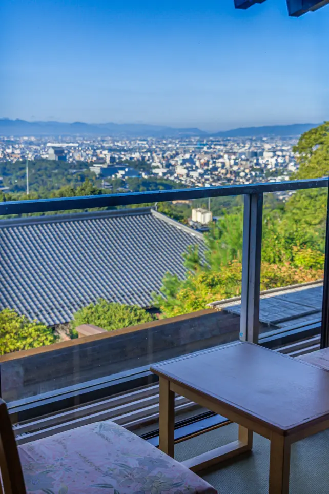 窓際の客室から夕暮れ時に見た景色。手前に木製の椅子と小さなテーブルがあり、窓越しには和風の屋根と、遠方に薄明かりに照らされた市街地が広がる。