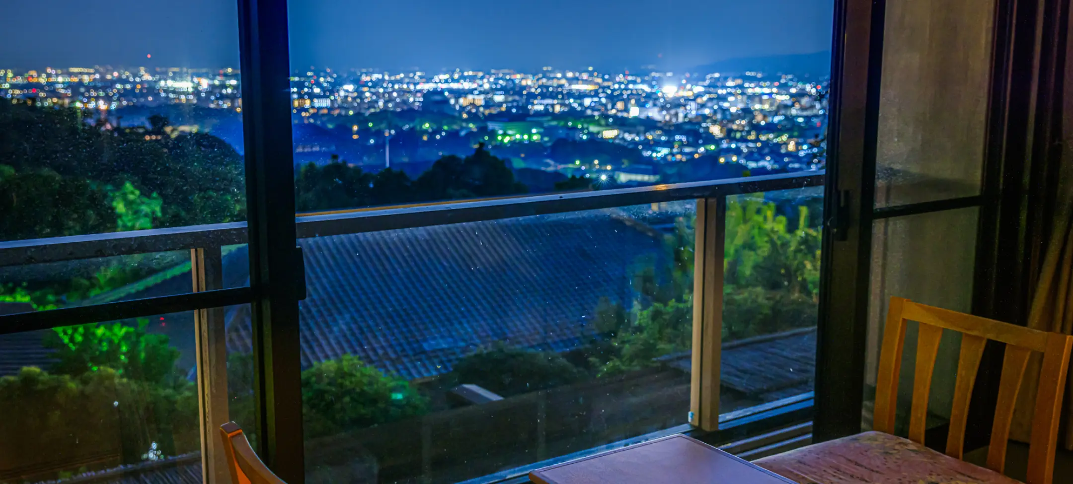 窓際の客室から夜間に見た景色。手前のテーブルと椅子は暗く、窓越しにはきらめく無数の灯りが広がる市街地の夜景が見える。