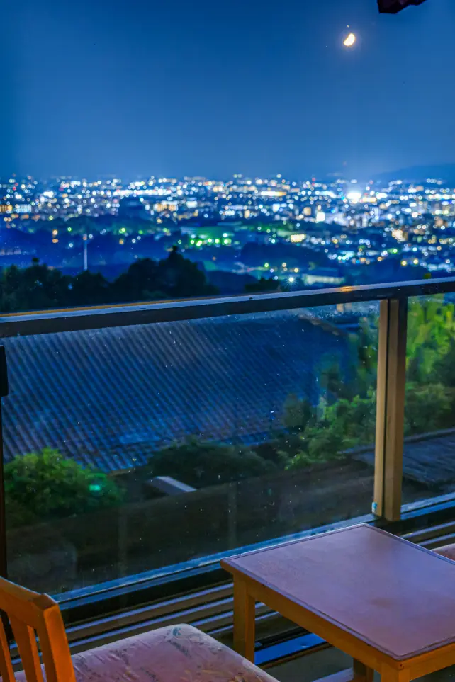 窓際の客室から夜間に見た景色。手前のテーブルと椅子は暗く、窓越しにはきらめく無数の灯りが広がる市街地の夜景が見える。