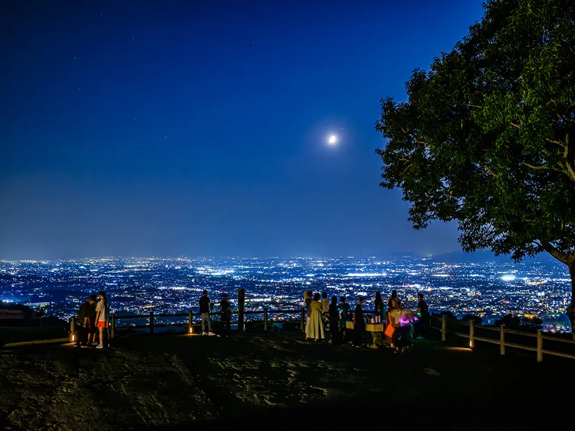 若草山から見下ろす奈良の夜景。満月が輝く夜空の下、展望台には複数の観光客が立ち、眼下に広がる街の光を眺めている。
