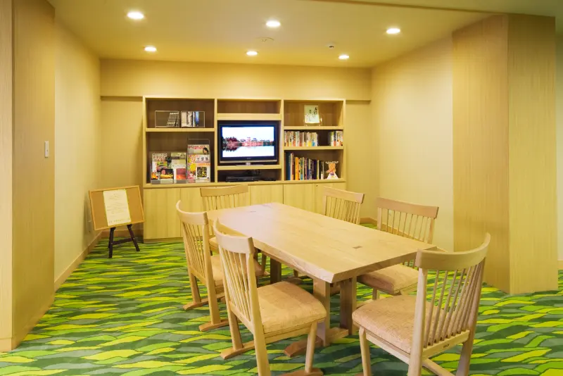 テレビと本棚が設置された一室に、大きな木製のテーブルと6脚の椅子が置かれている写真。