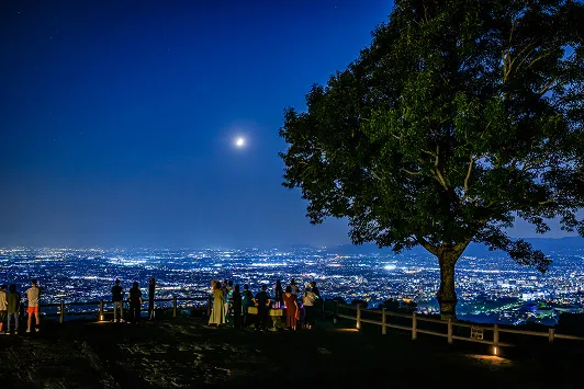 若草山から見下ろす奈良の夜景。満月が輝く夜空の下、展望台には複数の観光客が立ち、眼下に広がる街の光を眺めている。