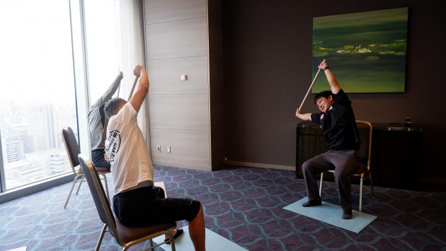 Image: A person stretching with their arms outstretched while sitting in a chair.