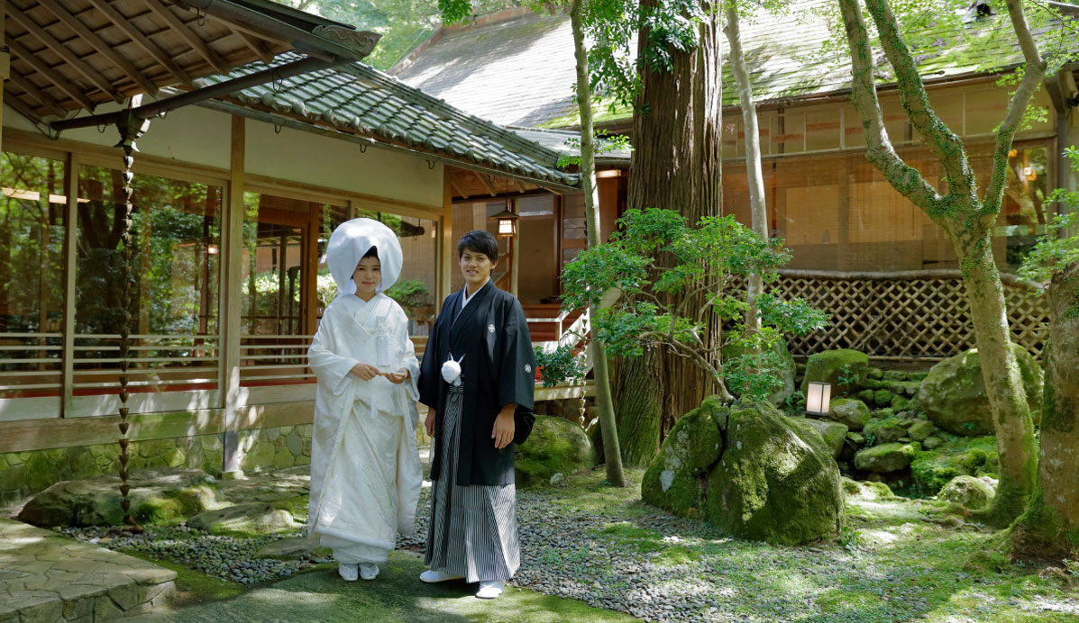 写真：日本庭園の中で結婚式の記念写真を撮影している新郎新婦。新婦は白無垢を着用し、新郎は黒の羽織袴を着用している。背景には木々や苔むした庭石、伝統的な日本家屋がある。